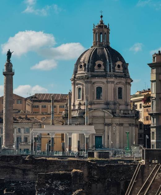 A large building with a dome on top and a tall column. The building is surrounded by other buildings and a street